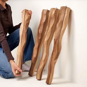 May include: Four tall, light brown wooden furniture legs with a distinct, hand-carved wavy or organic silhouette. Each leg features a smooth, polished finish with visible wood grain patterns and a small metal threaded bolt protruding from the top.