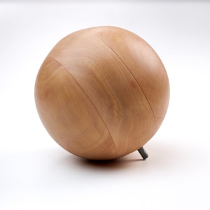 Puede incluir: Objeto esférico de madera clara con acabado marrón claro. La esfera tiene dos ranuras diagonales y descansa sobre una pequeña pata de metal oscuro. La veta de la madera es visible, sobre fondo blanco.