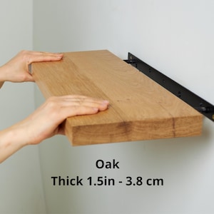 Pode incluir: Uma prateleira de carvalho castanho claro, com 3,8 cm de espessura, é suportada por um suporte de metal preto. A prateleira é retangular com grão de madeira visível. A palavra "Oak" está impressa na prateleira.