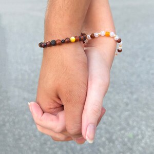 May include: Two people holding hands, each wearing a beaded bracelet. The bracelet on the left hand is made of brown, black, and yellow beads. The bracelet on the right hand is made of white and yellow beads.