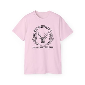 May include: A pale pink t-shirt with a distressed graphic. The graphic features a deer head inside a circle with the text "Brownville's Food Pantry for Deer" in a vintage style. The shirt is a crew neck style.