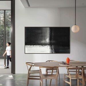 May include: A black and white abstract painting hangs on a white wall in a modern dining room. The painting features a horizontal black stripe across the top and a white stripe across the bottom, with a gray area in the middle. The painting is framed in black.