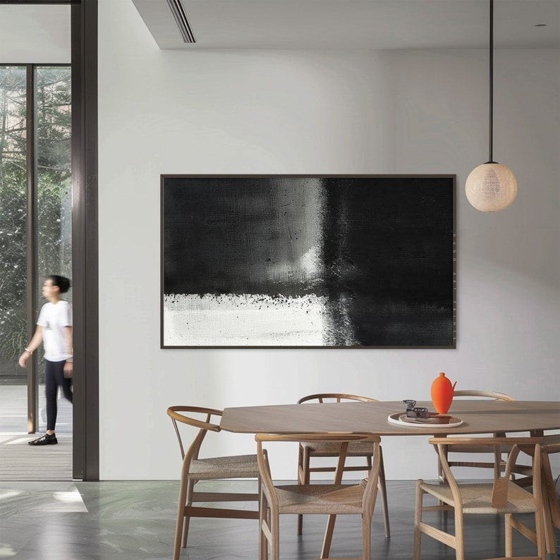 May include: A black and white abstract painting hangs on a white wall in a modern dining room. The painting features a horizontal black stripe across the top and a white stripe across the bottom, with a gray area in the middle. The painting is framed in black.