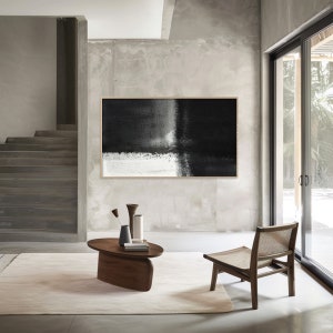 May include: A black and white abstract painting hangs on a wall in a modern living room. The painting is framed and features a large black area with a white stripe running through it. The room is decorated with a wooden coffee table and a chair.