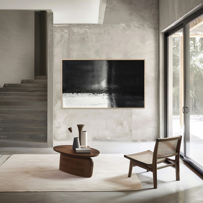 May include: A black and white abstract painting hangs on a wall in a modern living room. The painting is framed and features a large black area with a white stripe running through it. The room is decorated with a wooden coffee table and a chair.