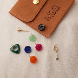 May include: A collection of colorful charms and a brown leather pouch with the text "MOD" and a logo. The charms include a green heart, a blue button, a red button, an orange button, a gold key, and a gold pin.