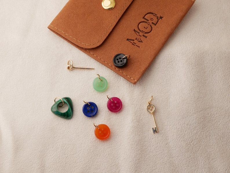 May include: A collection of colorful charms and a brown leather pouch with the text "MOD" and a logo. The charms include a green heart, a blue button, a red button, an orange button, a gold key, and a gold pin.