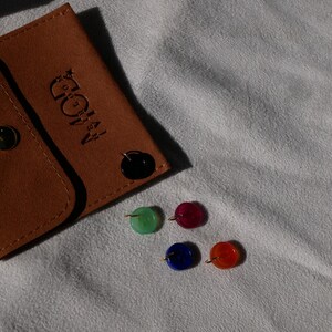 May include: A brown leather pouch with a gold snap closure and the embossed text "A MOD". Four colorful, round stud earrings in green, red, blue, and orange are displayed next to the pouch.
