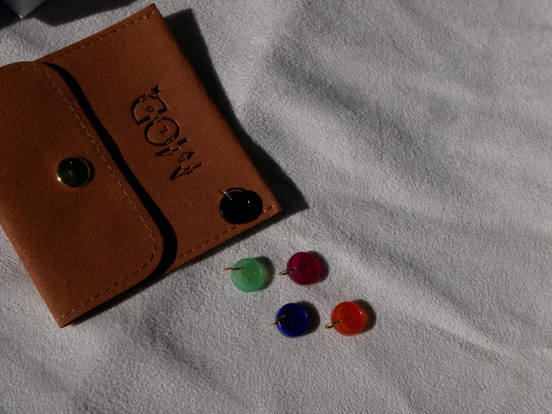 May include: A brown leather pouch with a gold snap closure and the embossed text "A MOD". Four colorful, round stud earrings in green, red, blue, and orange are displayed next to the pouch.