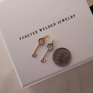 May include: Two key-shaped pendants, one gold-toned and one silver-toned, rest on a white box with the text "FOREVER WELDED JEWELRY". A US dime is next to the pendants for size comparison. The pendants have a detailed key design.