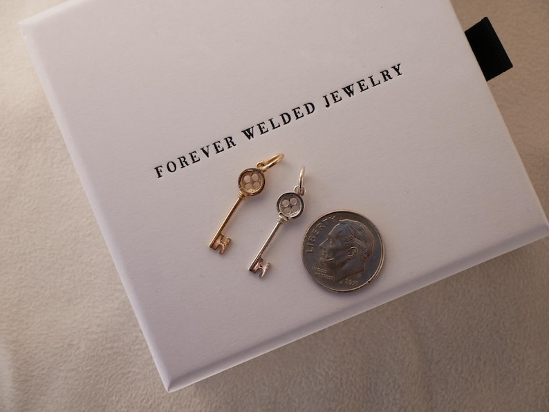May include: Two key-shaped pendants, one gold-toned and one silver-toned, rest on a white box with the text "FOREVER WELDED JEWELRY". A US dime is next to the pendants for size comparison. The pendants have a detailed key design.