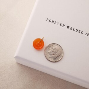 May include: An orange button pendant with a gold bail next to a dime. The pendant is round with four holes and a shiny finish. The dime is silver and shows a profile. The text "FOREVER WELDED JEWELRY" is printed on the white surface.