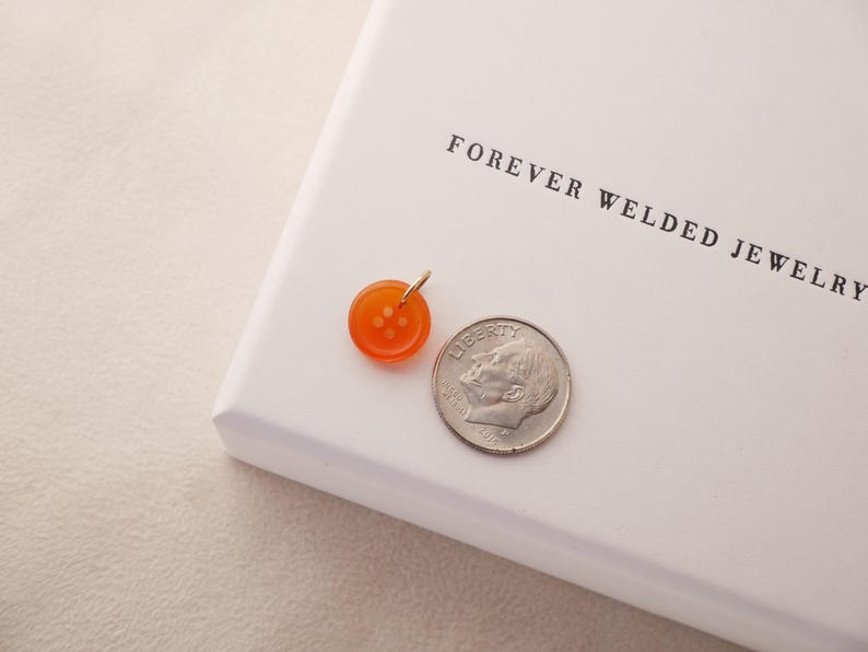 May include: An orange button pendant with a gold bail next to a dime. The pendant is round with four holes and a shiny finish. The dime is silver and shows a profile. The text "FOREVER WELDED JEWELRY" is printed on the white surface.