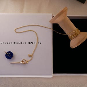 May include: A white box with the text "FOREVER WELDED JEWELRY" contains a gold necklace, a blue button charm, and a gold bar charm. A wooden spool holds a gold bracelet. The box is open, revealing a black interior.