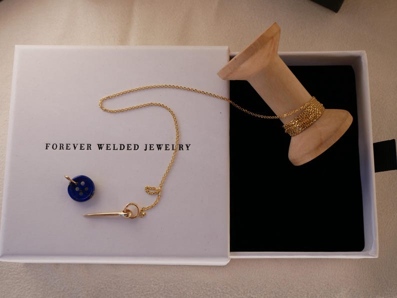 May include: A white box with the text "FOREVER WELDED JEWELRY" contains a gold necklace, a blue button charm, and a gold bar charm. A wooden spool holds a gold bracelet. The box is open, revealing a black interior.