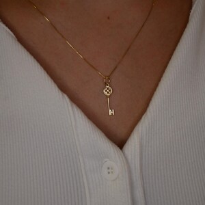 May include: A delicate gold necklace featuring a key pendant. The key has a detailed design on the head and a simple, straight shaft. The necklace is worn against a white collared shirt.
