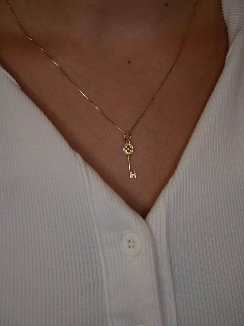 May include: A delicate gold necklace featuring a key pendant. The key has a detailed design on the head and a simple, straight shaft. The necklace is worn against a white collared shirt.