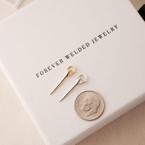 May include: Two delicate metal hair pins, one gold and one silver, rest on a white box with the text "FOREVER WELDED JEWELRY". A US dime is next to the pins for size comparison. The pins have a simple, elegant design.