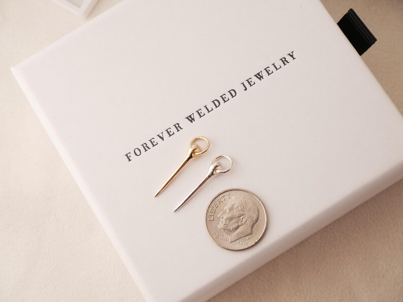 May include: Two delicate metal hair pins, one gold and one silver, rest on a white box with the text "FOREVER WELDED JEWELRY". A US dime is next to the pins for size comparison. The pins have a simple, elegant design.