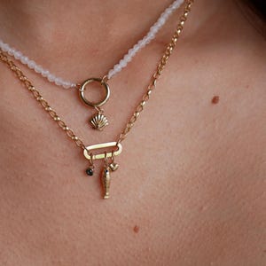 May include: A gold chain necklace with a charm of a seashell, a heart, a fish, and a small black stone. The necklace is layered over a white beaded necklace.