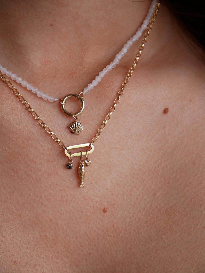 May include: A gold chain necklace with a charm of a seashell, a heart, a fish, and a small black stone. The necklace is layered over a white beaded necklace.