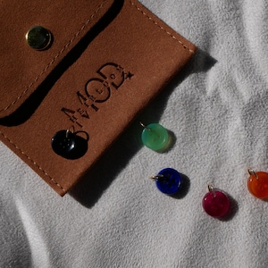 May include: A brown suede pouch with the text "4 MOD" and several colorful gemstone pendants. The pendants are in shades of black, green, blue, red, and orange. The pouch has a gold button closure.
