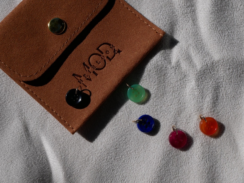 May include: A brown suede pouch with the text "4 MOD" and several colorful gemstone pendants. The pendants are in shades of black, green, blue, red, and orange. The pouch has a gold button closure.