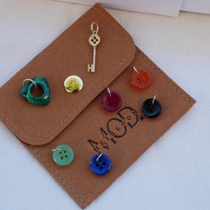 May include: A collection of jewelry charms and buttons displayed on a brown felt pouch. Charms include a gold key, a green stone, and a gold button. Buttons are red, black, blue, and green. The pouch has the text "44 MOD" printed on it.