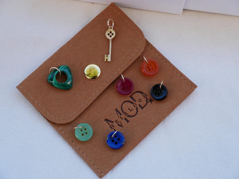May include: A collection of jewelry charms and buttons displayed on a brown felt pouch. Charms include a gold key, a green stone, and a gold button. Buttons are red, black, blue, and green. The pouch has the text "44 MOD" printed on it.