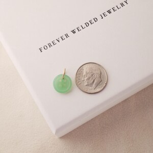 May include: A light green button pendant with a gold bail next to a dime on a white surface. The pendant is round with four holes and a slightly raised design. The text "FOREVER WELDED JEWELRY" is printed on the white surface.