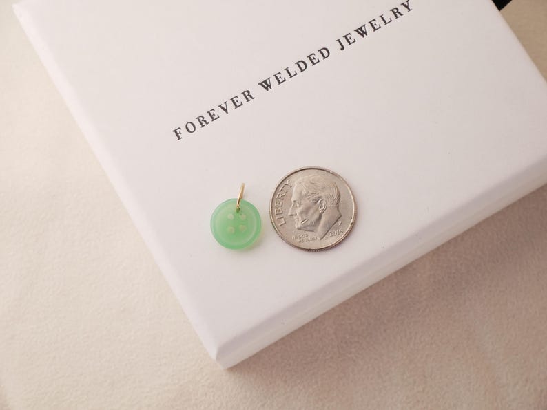 May include: A light green button pendant with a gold bail next to a dime on a white surface. The pendant is round with four holes and a slightly raised design. The text "FOREVER WELDED JEWELRY" is printed on the white surface.