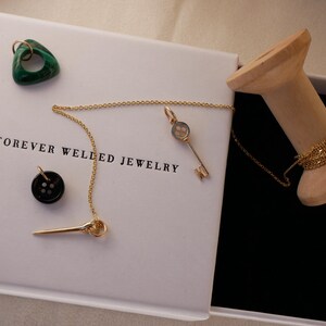 May include: A white box with the text "FOREVER WELLED JEWELRY" contains a gold necklace with a key pendant, a black button, and a green triangular stone. A wooden spool holds a gold chain. The box is open, revealing a black interior.