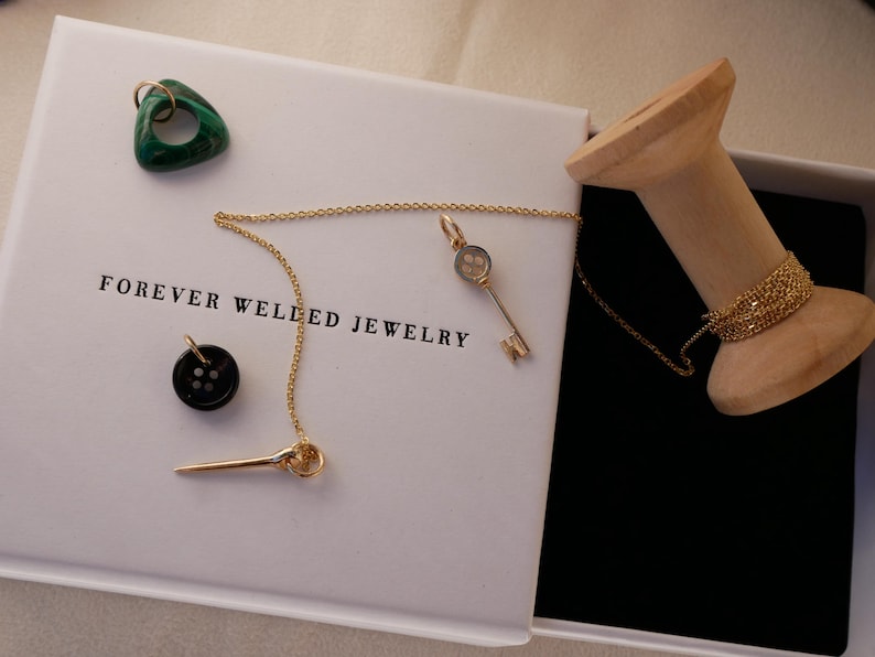 May include: A white box with the text "FOREVER WELLED JEWELRY" contains a gold necklace with a key pendant, a black button, and a green triangular stone. A wooden spool holds a gold chain. The box is open, revealing a black interior.