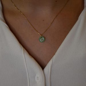 May include: A gold-colored chain necklace with a small, round pendant. The pendant is light green with a white floral design. The necklace is worn over a white ribbed shirt.
