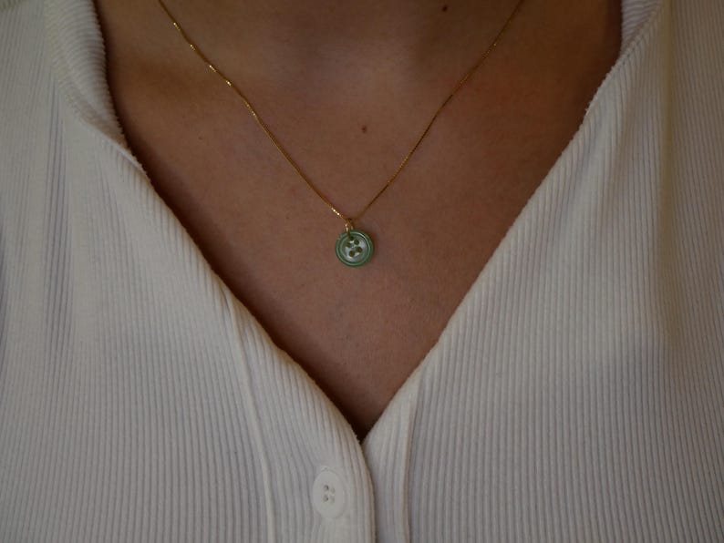 May include: A gold-colored chain necklace with a small, round pendant. The pendant is light green with a white floral design. The necklace is worn over a white ribbed shirt.