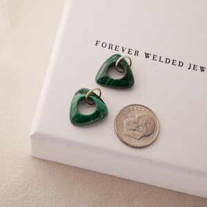 May include: Two triangular malachite pendants with silver and gold-colored hoops, next to a dime, on a white surface. The pendants have a dark green color with lighter green veining. The text "FOREVER WELDED JEWELRY" is printed on the surface.