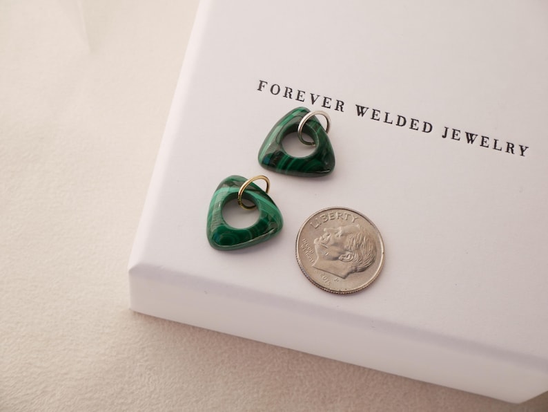 May include: Two triangular malachite pendants with silver and gold-colored hoops, next to a dime, on a white surface. The pendants have a dark green color with lighter green veining. The text "FOREVER WELDED JEWELRY" is printed on the surface.