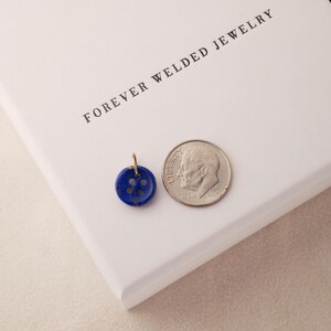 May include: A small, round, dark blue button pendant with four holes, next to a dime. The pendant has a gold-colored loop for a chain. The text "FOREVER WELDED JEWELRY" is printed on a white surface.
