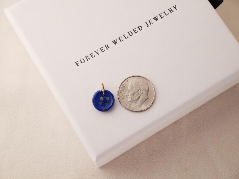 May include: A small, round, dark blue button pendant with four holes, next to a dime. The pendant has a gold-colored loop for a chain. The text "FOREVER WELDED JEWELRY" is printed on a white surface.