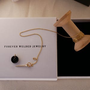 May include: An open white box with the text "FOREVER WELDED JEWELRY" contains a gold necklace, a black pendant, and a gold pin. A wooden spool holds a gold chain. The box has a black velvet interior.
