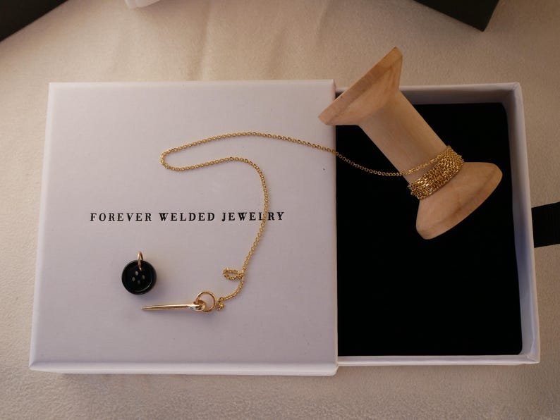 May include: An open white box with the text "FOREVER WELDED JEWELRY" contains a gold necklace, a black pendant, and a gold pin. A wooden spool holds a gold chain. The box has a black velvet interior.
