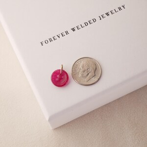 May include: A small, round, fuchsia-colored pendant with a gold-colored bail next to a dime on a white surface. The pendant is approximately 0.5 inches in diameter. The text "FOREVER WELDED JEWELRY" is printed in black.