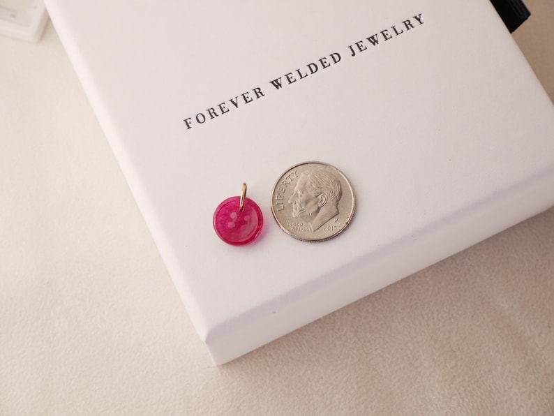May include: A small, round, fuchsia-colored pendant with a gold-colored bail next to a dime on a white surface. The pendant is approximately 0.5 inches in diameter. The text "FOREVER WELDED JEWELRY" is printed in black.