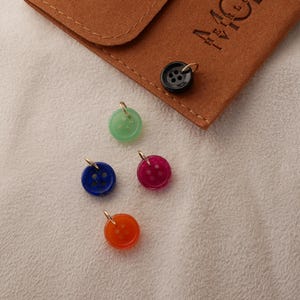 May include: A collection of colorful button charms with gold-tone loops. The charms are in shades of green, blue, red, and orange. A brown suede pouch with the letters "MC" is in the background. The buttons are on a white textured surface.