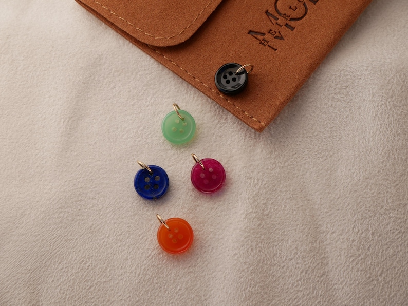 May include: A collection of colorful button charms with gold-tone loops. The charms are in shades of green, blue, red, and orange. A brown suede pouch with the letters "MC" is in the background. The buttons are on a white textured surface.