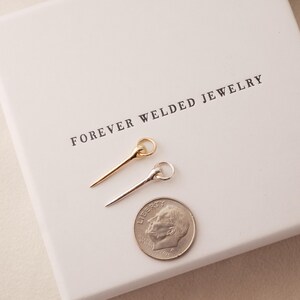 May include: Two delicate pin-shaped jewelry pieces, one gold and one silver, are displayed on a white box with the text "FOREVER WELDED JEWELRY". A US dime is placed next to the jewelry for size comparison. The jewelry has a ring at the top.