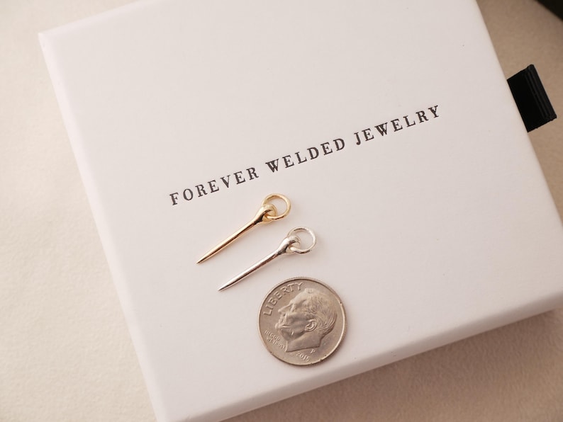 May include: Two delicate pin-shaped jewelry pieces, one gold and one silver, are displayed on a white box with the text "FOREVER WELDED JEWELRY". A US dime is placed next to the jewelry for size comparison. The jewelry has a ring at the top.
