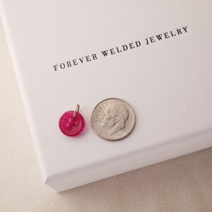 May include: A small, round, pink pendant with a gold-colored bail next to a dime. The pendant is made of a translucent material. The text "FOREVER WELDED JEWELRY" is printed on the white box in the background.
