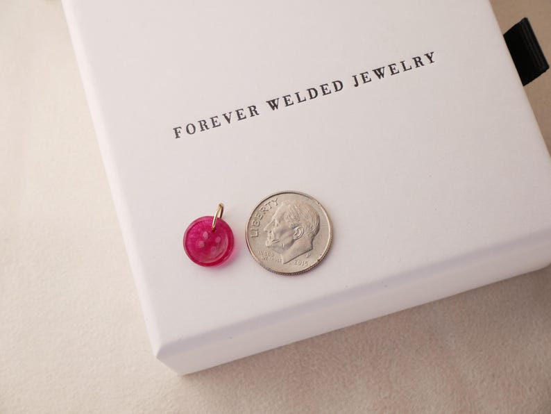 May include: A small, round, pink pendant with a gold-colored bail next to a dime. The pendant is made of a translucent material. The text "FOREVER WELDED JEWELRY" is printed on the white box in the background.