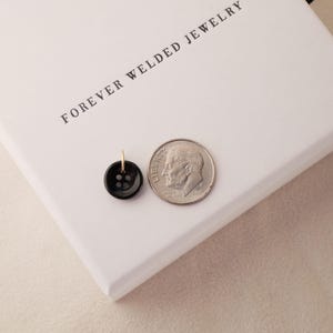May include: A black button pendant with a gold-colored bail next to a dime on a white box. The button has four holes and a shiny finish. The box has the words "FOREVER WELDED JEWELRY" printed on it.